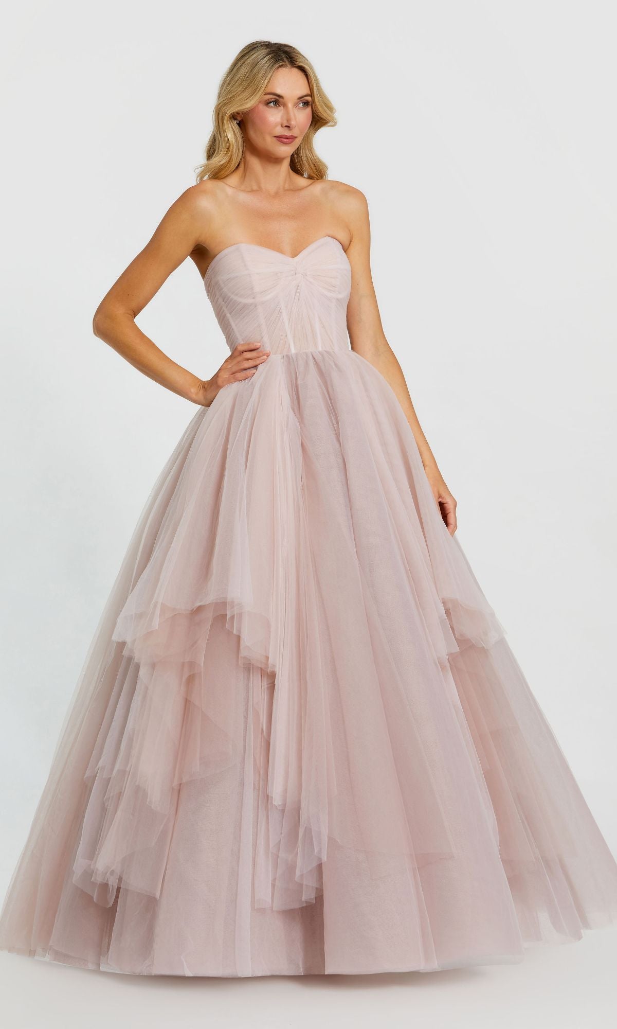 A woman models the Long Formal Dress 20772 by Mac Duggal, featuring a strapless ruched bodice and layered tulle ball gown skirt in light pink, standing against a plain white background.